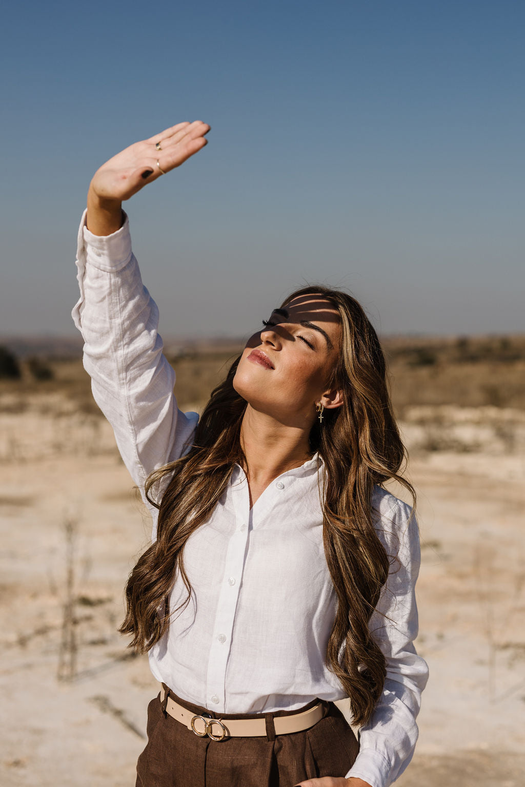 A woman outdoors in a desert landscape wearing  the Ivory 100% certified Irish linen in a long-sleeve button-up shirt, posing with her hand raised to shield her eyes from the sun.