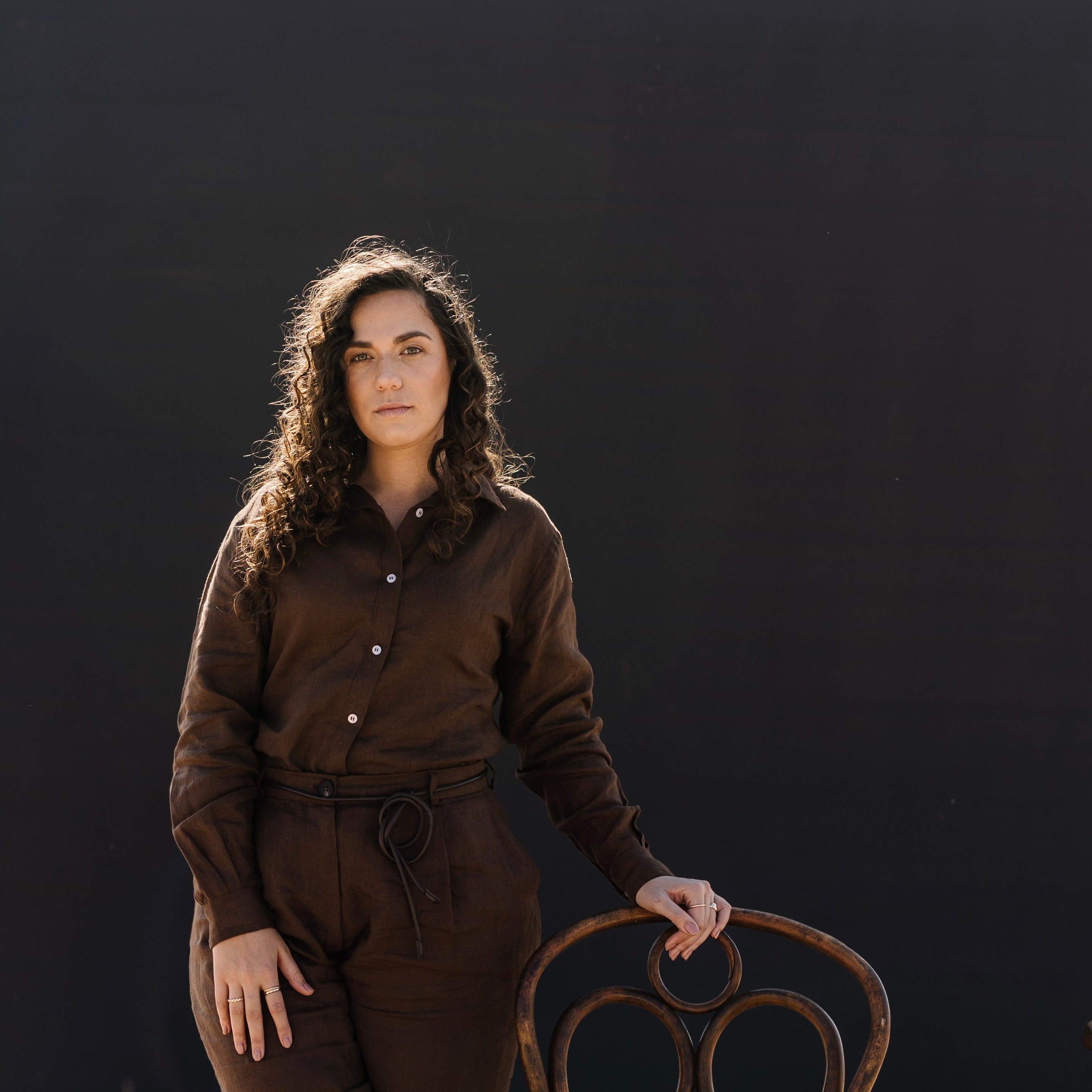 A woman wearing a matching Lowveld Umber 100% European brown linen button-up shirt and  Lowveld Umber 100% European brown linen pants , standing outdoors next to a wooden chair in front of a large black backdrop board.