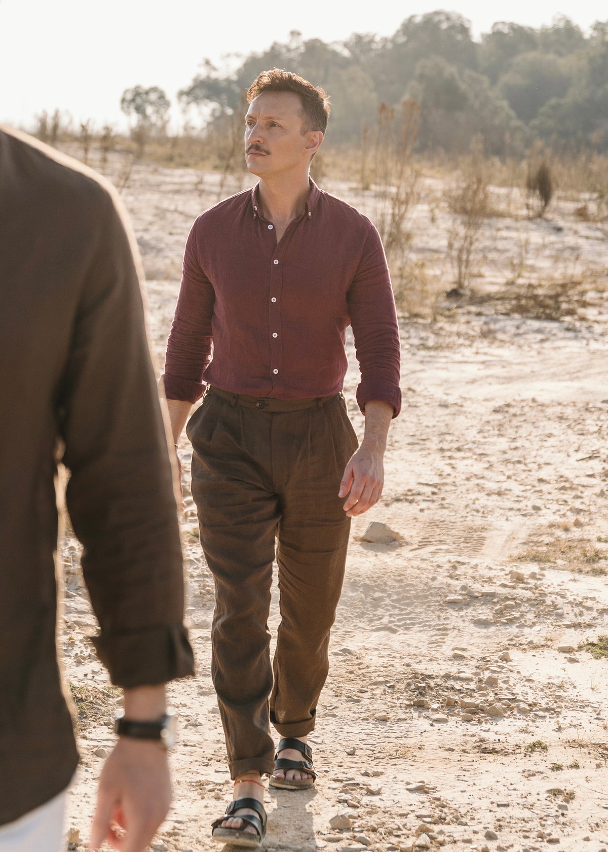 A man wearing  a Royal Karoo 100% certified Irish burgundy linen button-up shirt and Lowveld Umber 100% European dark brown linen trousers and sandals walks along a sandy path outdoors, with another person partially visible in the foreground.