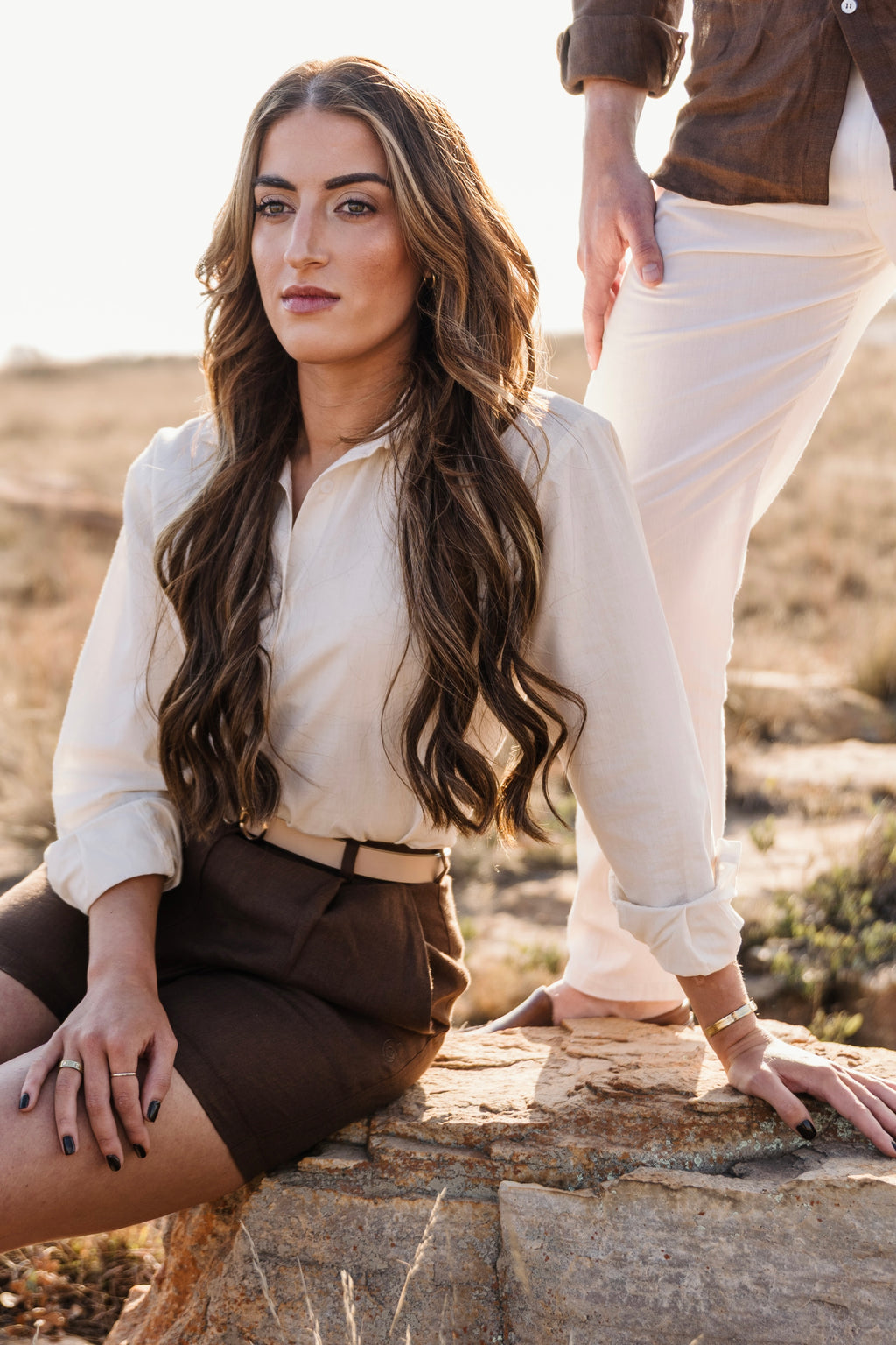 A woman with long wavy brown hair sitting outdoors on a rock in a dry grassy field, wearing a cream Tsitsikamma Mist 100% Egyptian linen button-down shirt tucked into a Lowveld Umber 100% European brown linen  trousers with a tan belt.