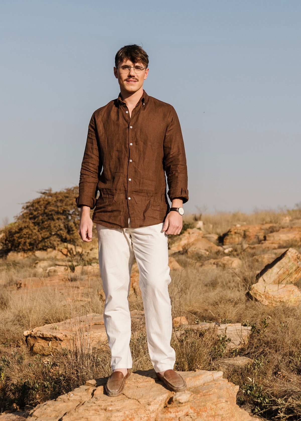 A man wearing a matching Lowveld Umber 100% European brown linen button-up shirt and cream Tsitsikamma Mist 100% Egyptian linen  trousers stands outdoors on a rocky, dry grassy landscape under a clear blue sky.