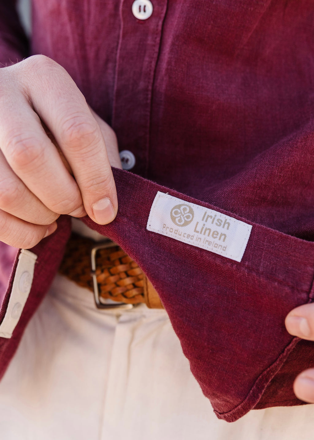 Close-up of a person holding open a Royal Karoo 100% certified burgandy linen shirt to show the inside label, which reads 'Irish Linen Produced in Ireland which is the Irish guild and certification