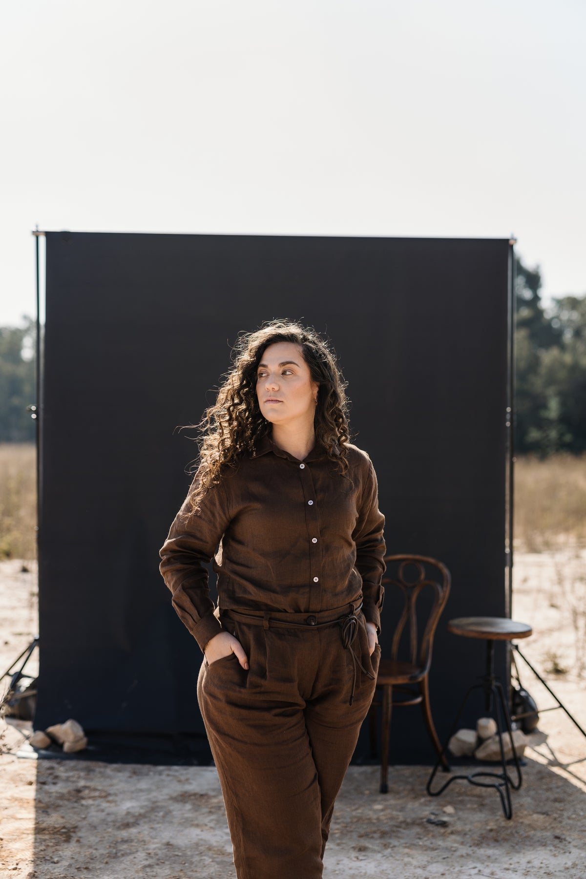A woman wearing a matching Lowveld Umber 100% European brown linen button-up shirt and  Lowveld Umber 100% European brown linen pants , standing outdoors  in front of a large black backdrop board.