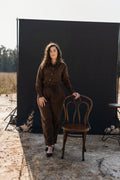 A woman wearing a matching Lowveld Umber 100% European brown linen button-up shirt and  Lowveld Umber 100% European brown linen pants , standing outdoors next to a wooden chair in front of a large black backdrop board.