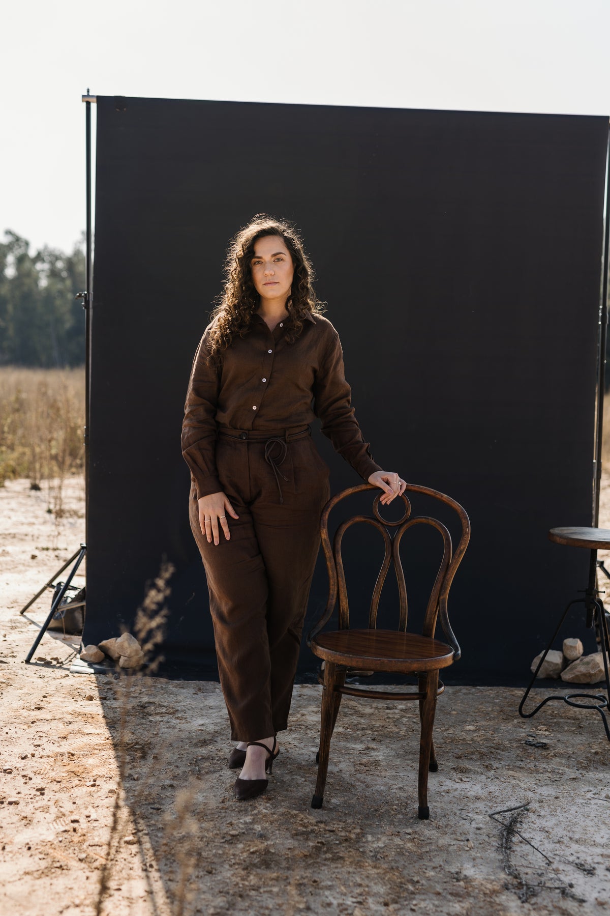 A woman wearing a matching Lowveld Umber 100% European brown linen button-up shirt and  Lowveld Umber 100% European brown linen pants , standing outdoors next to a wooden chair in front of a large black backdrop board.