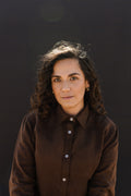 A woman with curly brown hair wearing a matching Lowveld Umber 100% European brown linen button-up shirt, photographed against a dark studio background.