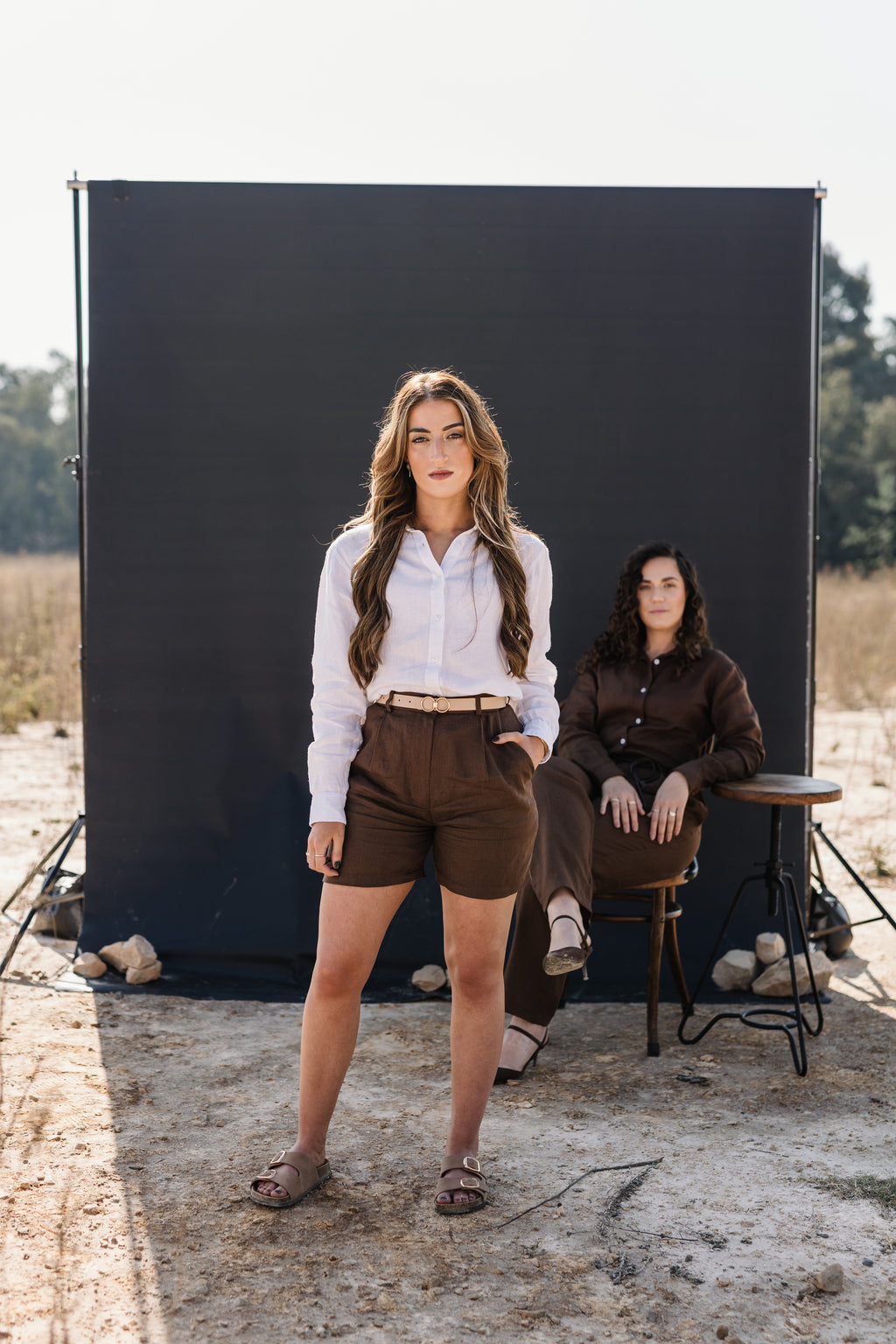 A woman stands in front of a large black photography backdrop outdoors on a dirt field, wearing a cream Tsitsikamma Mist 100% Egyptian linen button-up shirt and Lowveld Umber 100% European brown linen shorts. Another woman sits on a stool behind her to the right, wearing a Lowveld Umber 100% European linen set.