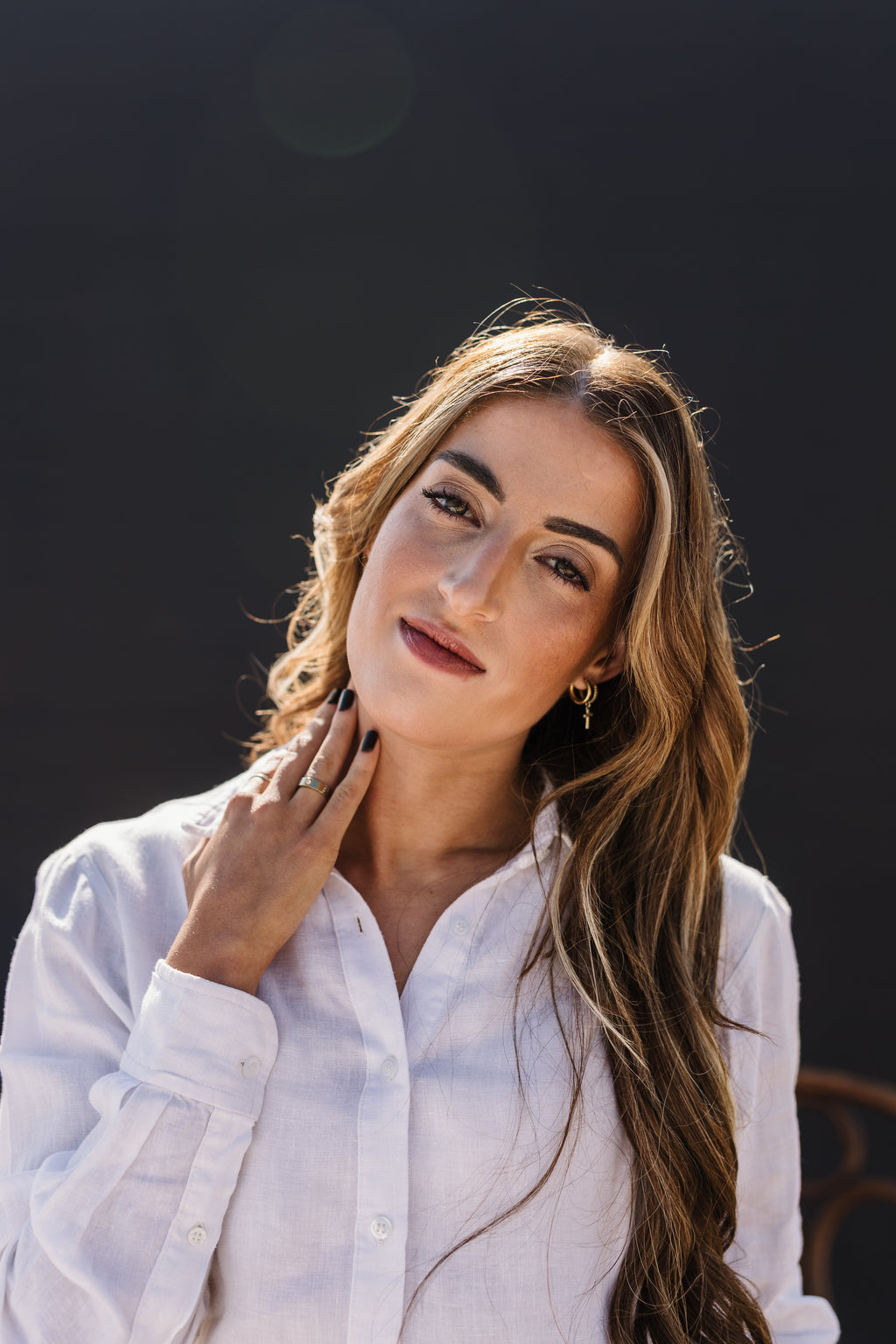 A woman with long wavy brown hair wearing the Ivory 100% certified Irish linen in a long-sleeve button-up shirt, posing against a dark, out-of-focus background.