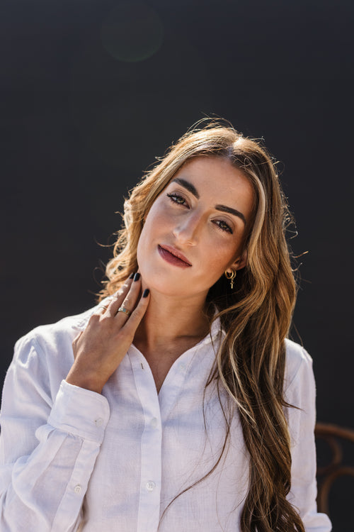 A woman with long wavy brown hair wearing the Ivory 100% certified Irish linen in a long-sleeve button-up shirt, posing against a dark, out-of-focus background.