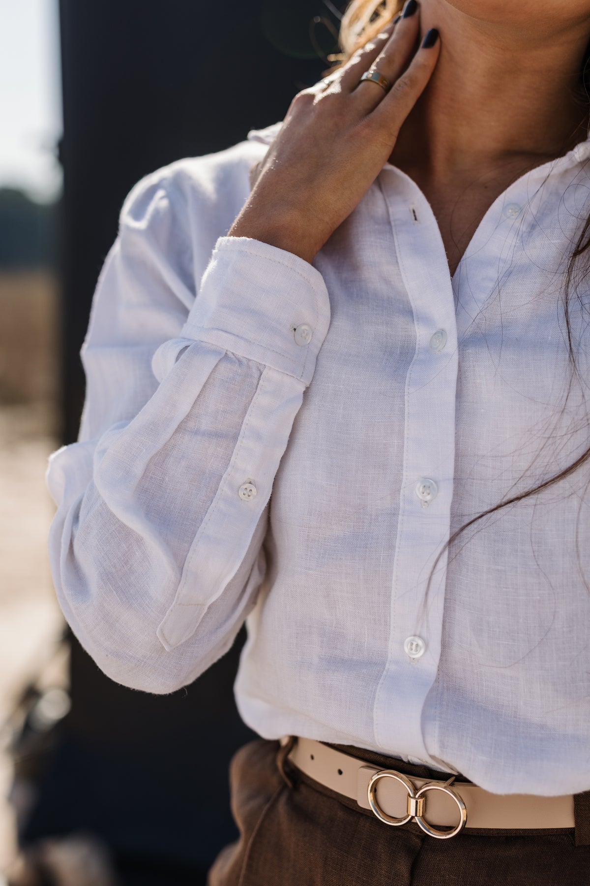 A close-up of a person wearing  the Ivory 100% certified Irish linen in a long-sleeve button-up shirt, posing against a dark, out-of-focus background white long-sleeve linen button-up shirt tucked into Lowveld Umber 100% European  brown linen trousers with a tan belt featuring double gold rings.