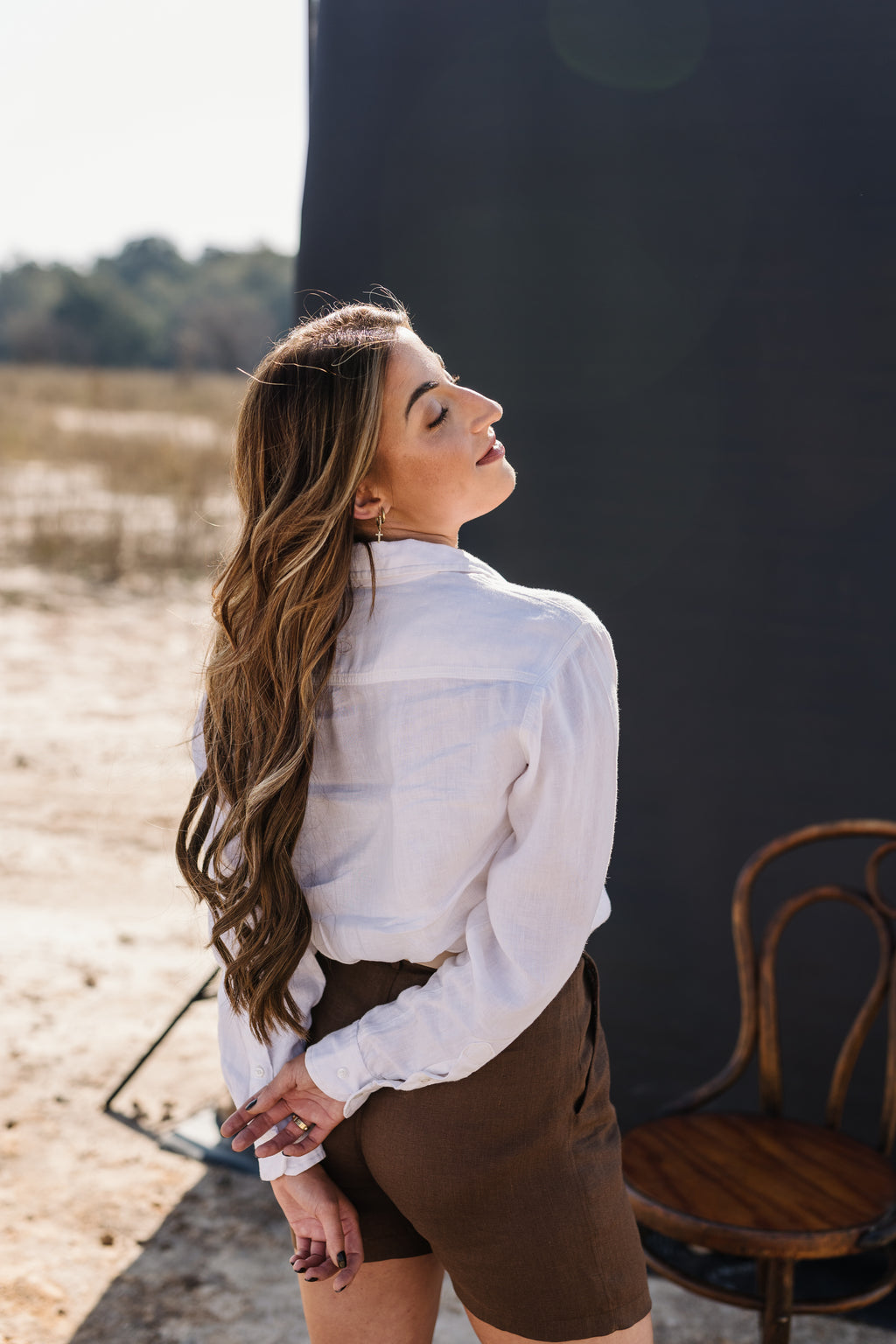 A woman with long wavy brown hair wearing the Ivory 100% certified Irish linen in a long-sleeve button-up shirt, posing against a dark, out-of-focus background.