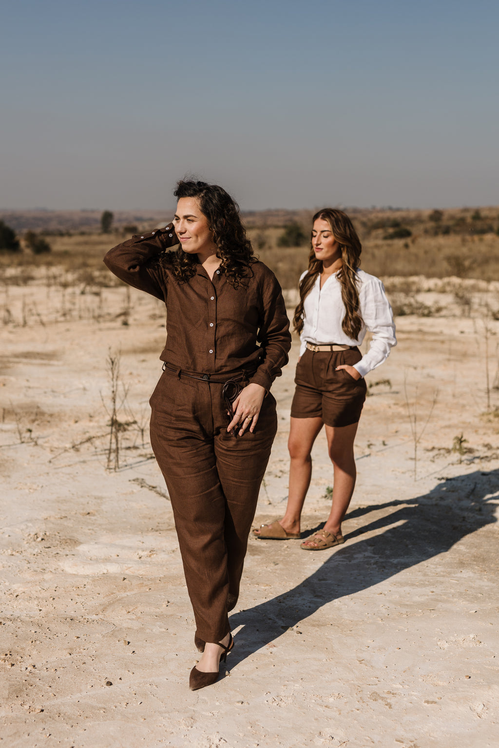 Two women walking outdoors in a dry, sandy landscape. The woman in the foreground wears a matching  Lowveld Umber 100% European brown linen button-up shirt and  Lowveld Umber 100% European brown linen pants, while the woman behind her wears a cream Tsitsikamma Mist 100% Egyptian linen  button-down shirt and Lowveld Umber 100% European brown linen shorts.