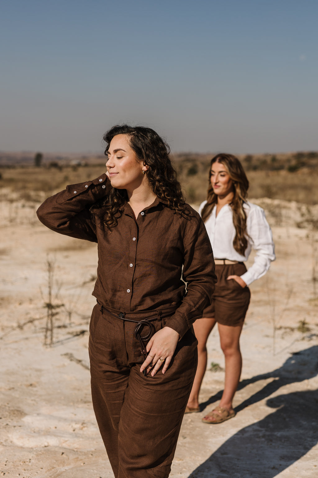 Two women walking outdoors in a dry, sandy landscape. The woman in the foreground is wearing a matching set of Lowveld Umber 100% European brown linen button-up shirt and  Lowveld Umber 100% European brown linen trousers set, while the woman behind her wears a cream Tsitsikamma Mist 100% Egyptian linen  button-down shirt  and  Lowveld Umber 100% European brown linen shorts.