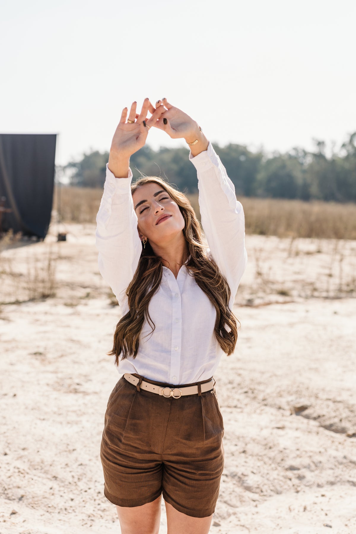 A woman stands outdoors in a sandy field wearing a cream 
Tsitsikamma Mist 100% Egyptian linen long-sleeve button-up shirt and Lowveld Umber 100% European brown linen shorts with a beige belt. She has her arms raised above her head in a relaxed pose under a bright sky.