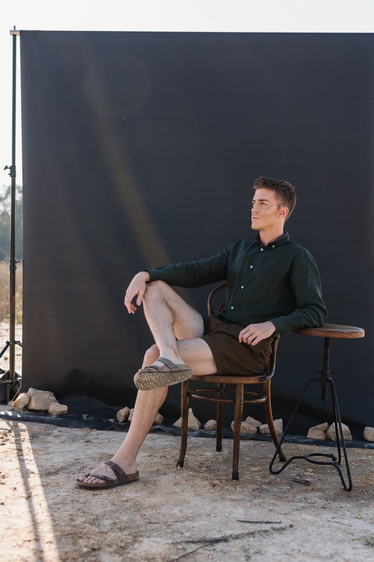 A man sits on a wooden stool in front of a black photography backdrop wearing a Drakensberk dawn 100% certified Irish dark green linen in a long-sleeve button-up shirt and Lowveld Umber 100% European  brown linen shorts 