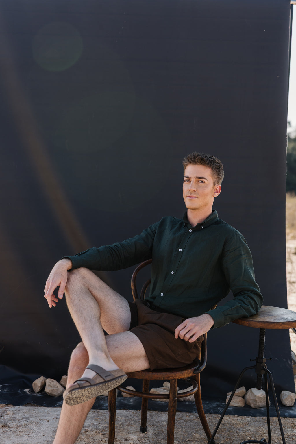 A man wearing a Drakensberk dawn 100% certified Irish dark green linen in a long-sleeve button-up shirt and Lowveld Umber 100% European  brown linen shorts sitting in front of a black photography backdrop in an outdoor studio setting 
