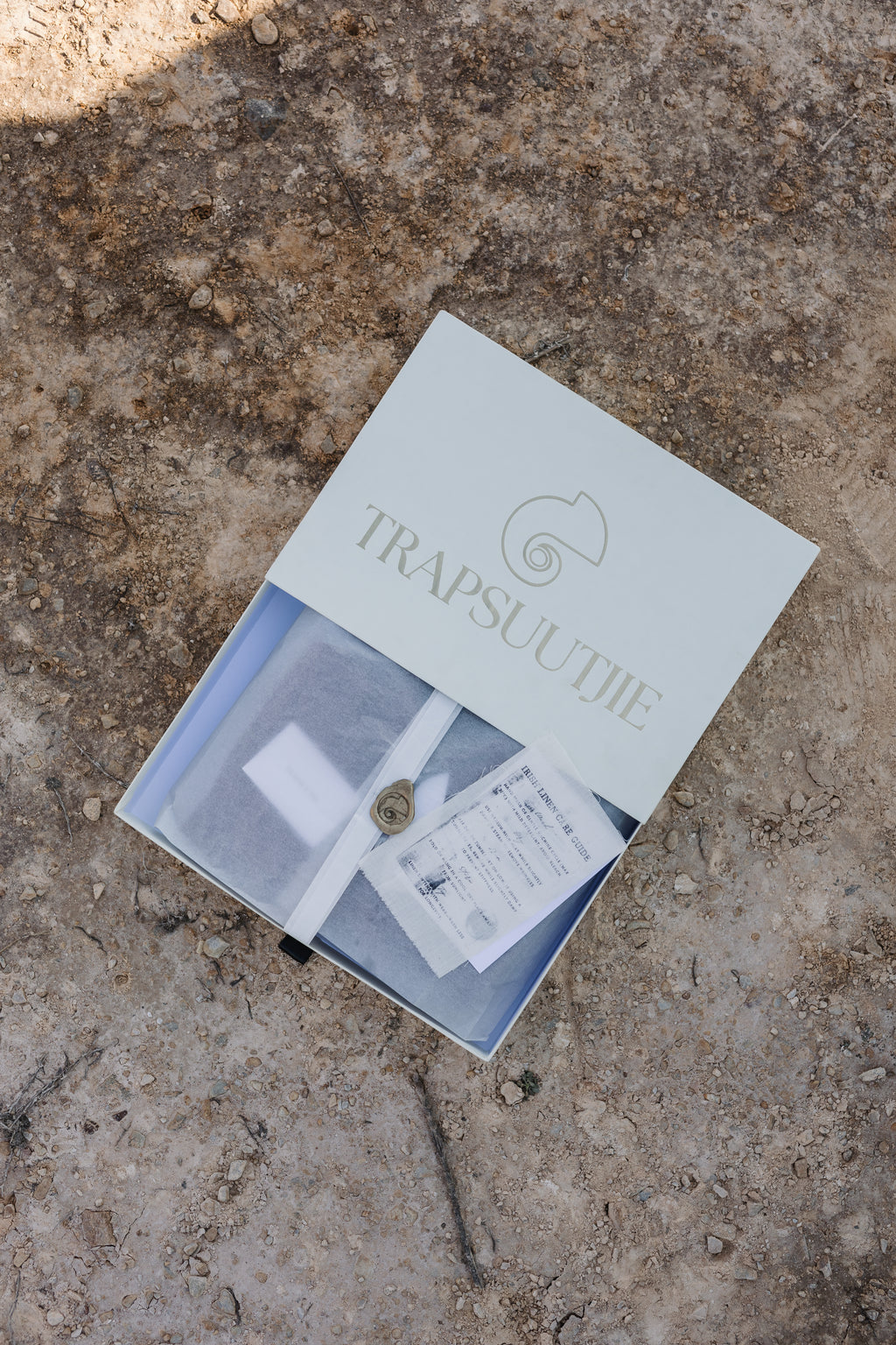 A cream gift box for Trapsuutjie, containing a wrapped Trapsuutjie linen shirt sealed with a browns wax stamp, placed on a textured sandy surface.