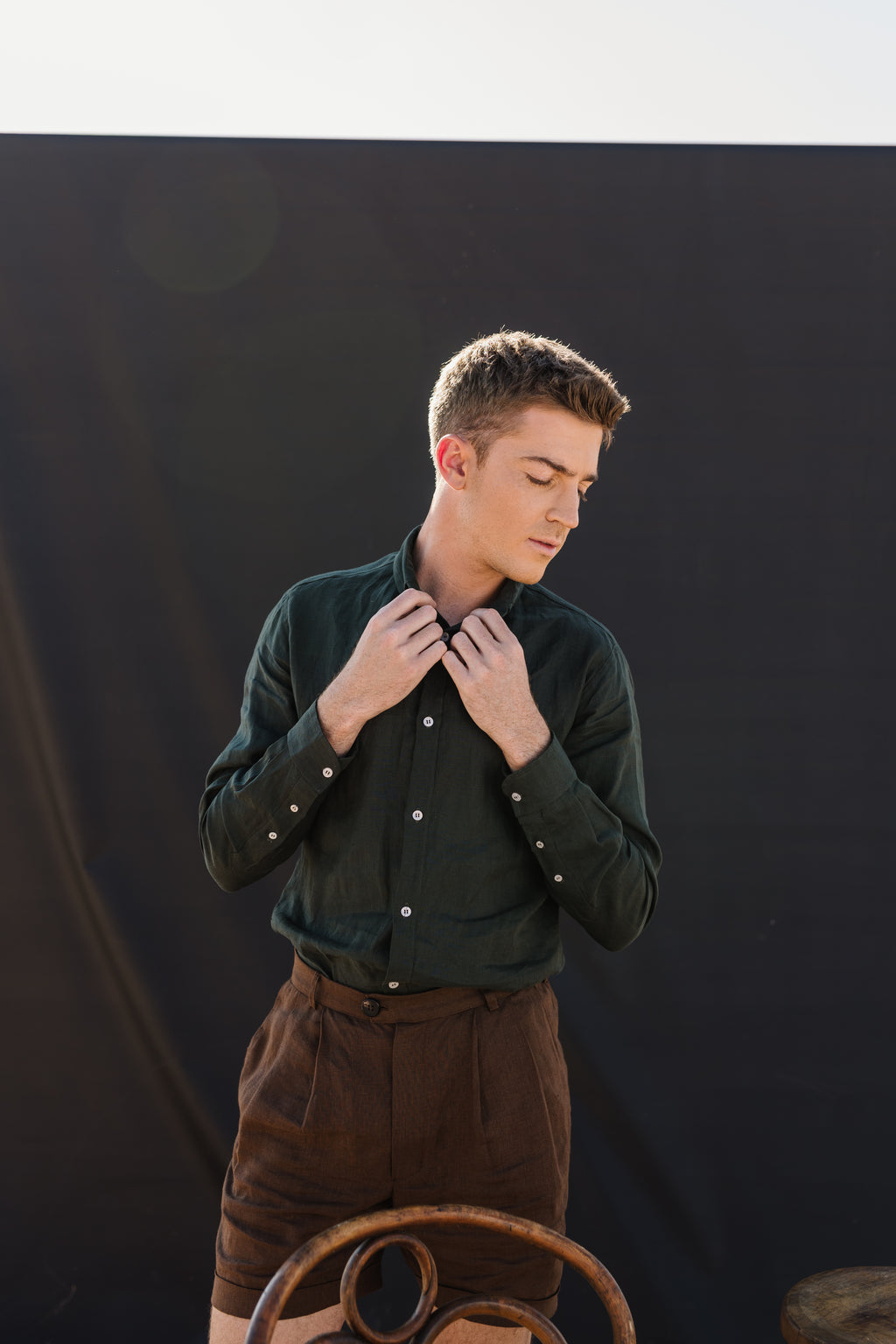 
A man wearing a Drakensberg dawn 100% certified Irish dark green linen in a long-sleeve button-up shirt and Lowveld Umber 100% European  brown linen shorts walks in front of a black photography backdrop in an outdoor studio setting with a wooden chair and stool.