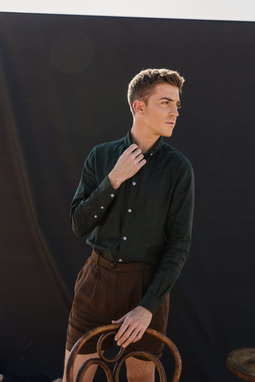 A man wearing a Drakensberk dawn 100% certified Irish dark green linen in a long-sleeve button-up shirt and Lowveld Umber 100% European  brown linen shorts, in front of a black photography backdrop in an outdoor studio setting with a wooden chair.