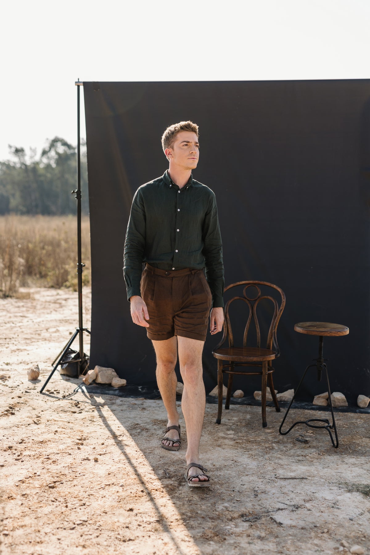 A man wearing a Drakensberk dawn 100% certified Irish dark green linen in a long-sleeve button-up shirt and Lowveld Umber 100% European  brown linen shorts walks in front of a black photography backdrop in an outdoor studio setting with a wooden chair and stool.
