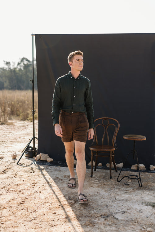 A man wearing a Drakensberk dawn 100% certified Irish dark green linen in a long-sleeve button-up shirt and Lowveld Umber 100% European  brown linen shorts walks in front of a black photography backdrop in an outdoor studio setting with a wooden chair and stool.