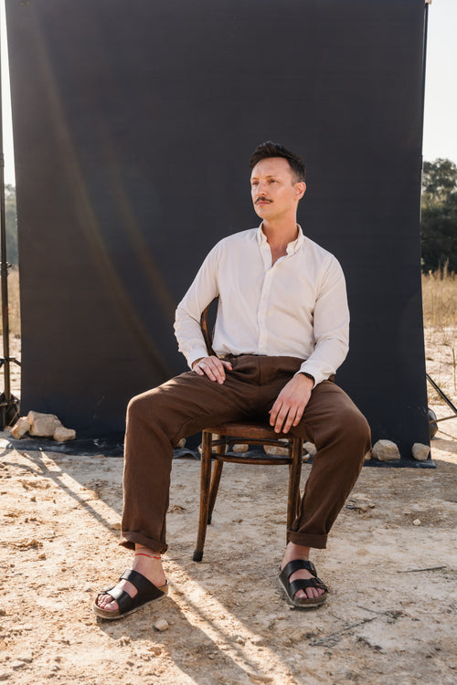 A man with a mustache wearing a cream Tsitsikamma Mist 100% Egyptian linen  button-down shirt and Lowveld Umber brown 100% linen trousers, sitting on a wooden chair against a dark backdrop.