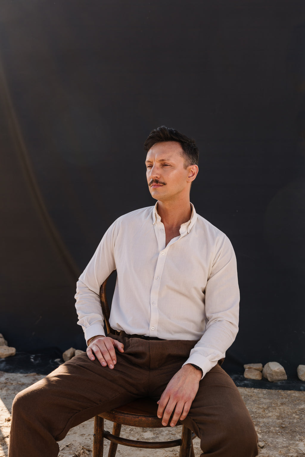 A man with a mustache wearing a cream Tsitsikamma Mist 100% Egyptian linen  button-down shirt and Lowveld Umber brown trousers, sitting on a wooden chair against a dark backdrop.