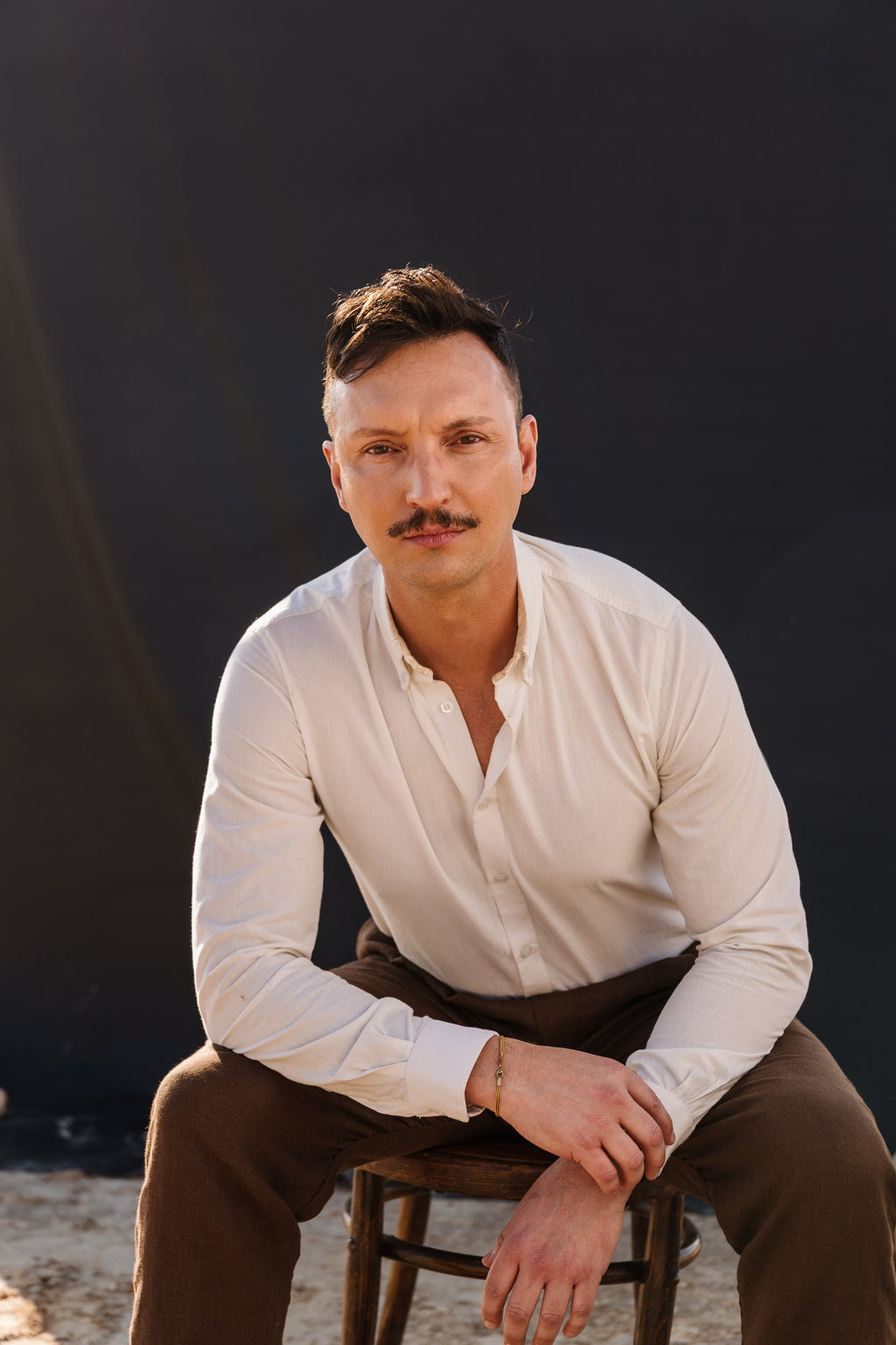 A man with a mustache wearing a cream Tsitsikamma Mist 100% Egyptian linen  button-down shirt and Lowveld Umber brown trousers, sitting on a wooden chair against a dark backdrop.