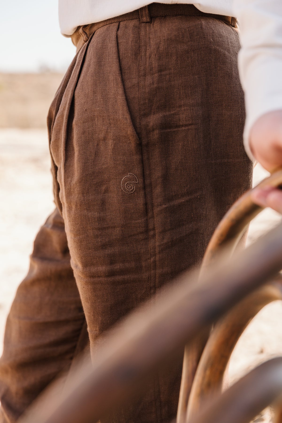 Close-up of a person wearing Lowveld Umber 100% European brown linen trousers in an outdoor sandy setting, with a blurred wooden chair in the foreground.