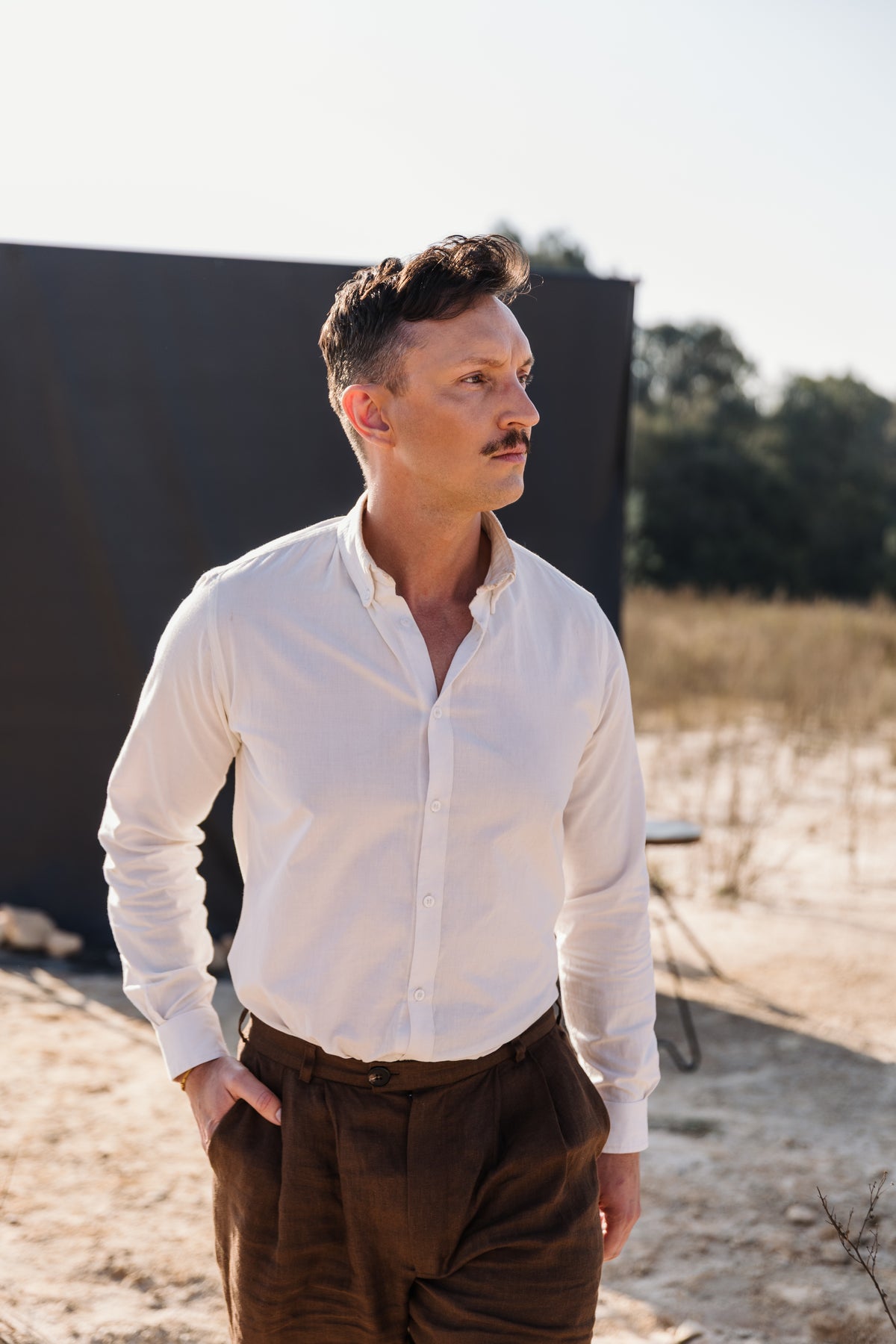A man with a mustache wearing a cream Tsitsikamma Mist 100% Egyptian linen  button-down shirt and Lowveld Umber brown trousers.