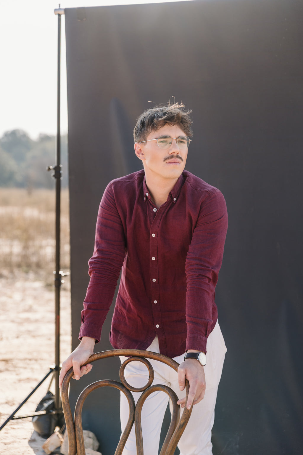 A man with glasses and a moustache sits on a wooden stool, wearing a Royal Karoo burgundy 100% certified Irish linen in a long-sleeve button-up shirt and and a cream Tsitsikamma Mist 100% trousers, set against a dark background