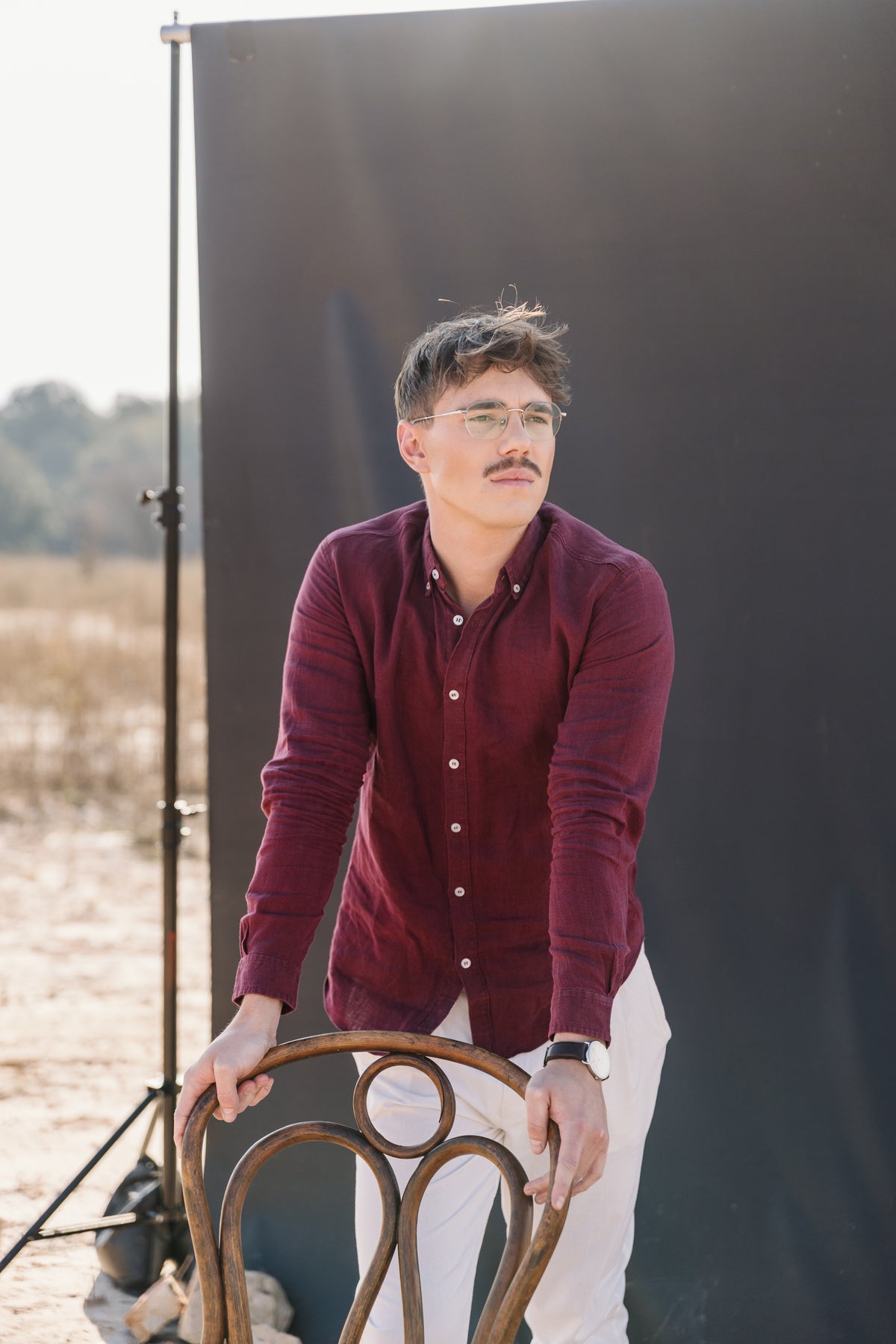 A man with glasses and a moustache sits on a wooden stool, wearing a Royal Karoo burgundy 100% certified Irish linen in a long-sleeve button-up shirt and and a cream Tsitsikamma Mist 100% trousers, set against a dark background