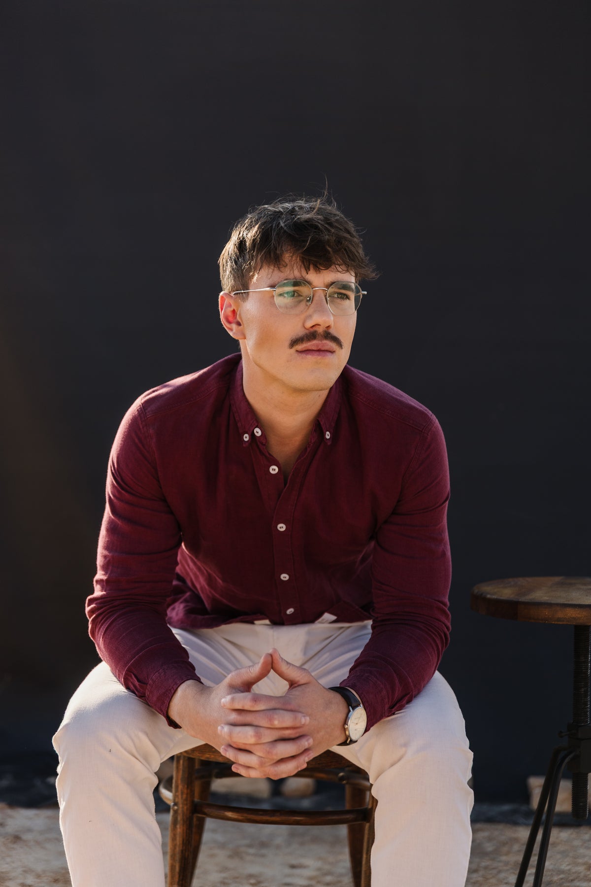 A man with glasses and a moustache sits on a wooden stool, wearing a burgundy 100% certified Irish  linen in a long-sleeve button-up shirt and a cream Tsitsikamma Mist 100% linen trousers, set against a dark background.