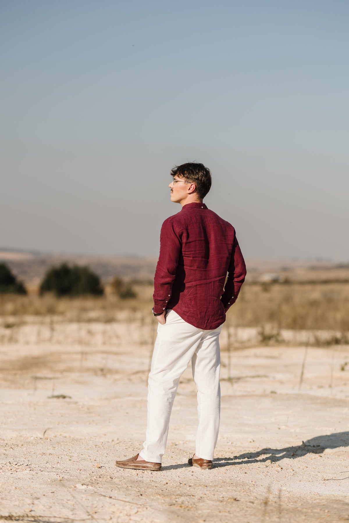 A man standing outdoors in a dry, sandy landscape, viewed from behind. A man with glasses and a moustache sits on a wooden stool, wearing a Royal Karoo burgundy 100% certified Irish linen in a long-sleeve button-up shirt and and a cream Tsitsikamma Mist 100% trousers, set against a dark background and brown shoes.
