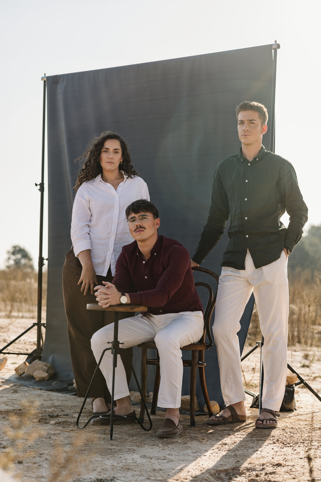 Three people posing outdoors in front of a large black photography backdrop stand. The three models are wearing the three shades of 100% certified Irish Linen, the two men are wearing cream Tsitsikamma Mist 100% Egyptian linen pants and woman is wearing a Lowveld Umber 100% European  brown linen trousers.