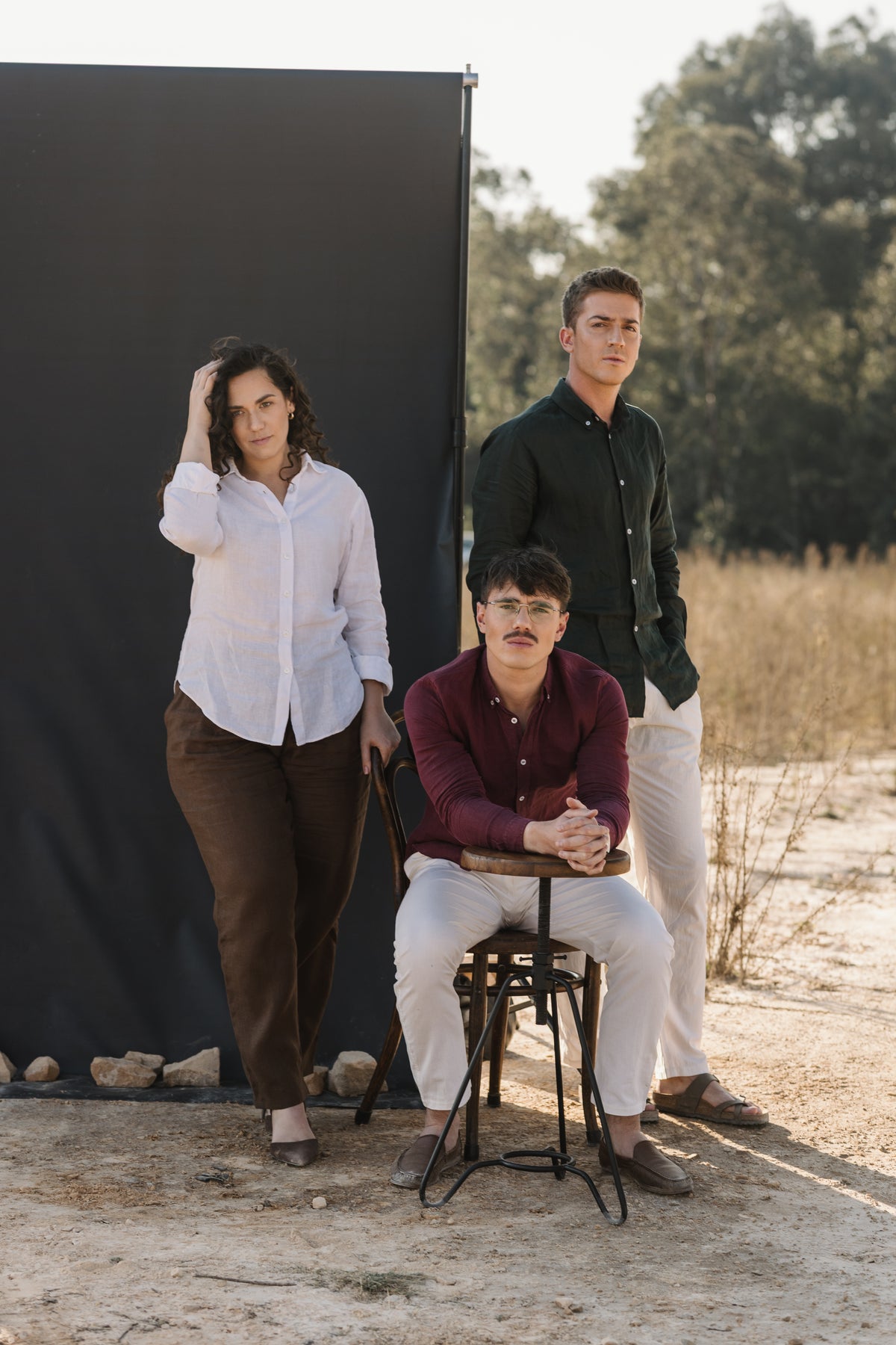 Three people posing outdoors in front of a large black photography backdrop stand. The three models are wearing the three shades of 100% certified Irish Linen, the two men are wearing cream Tsitsikamma Mist 100% Egyptian linen pants and woman is wearing a Lowveld Umber 100% European  brown linen trousers.