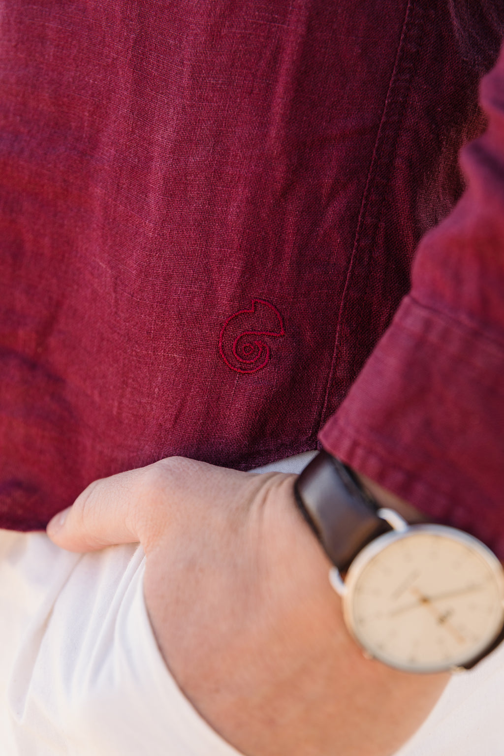 Close-up of a person wearing Royal Karoo burgundy 100% certified Irish linen  with a small embroidered Trapsuutjie logo, paired with a white cream Tsitsikamma Mist 100% Egyptian trousers and a dark brown leather strap watch.