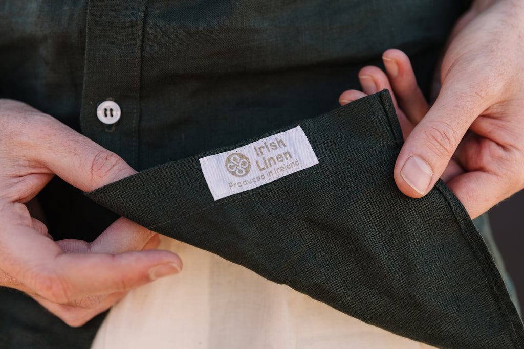 Close-up of a person holding open a Drakensberg dawn 100% certified Irish dark green linen shirt to show the inside label, which reads 'Irish Linen Produced in Ireland which is the Irish guild and certification 