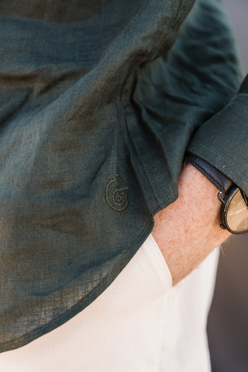 Close-up of a person wearing a Drakensberk dawn 100% certified Irish dark green linen small embroidered Trapsuutjie logo on the cuff, paired with a cream Tsitsikamma Mist 100% Egyptian linen pants and a watch.