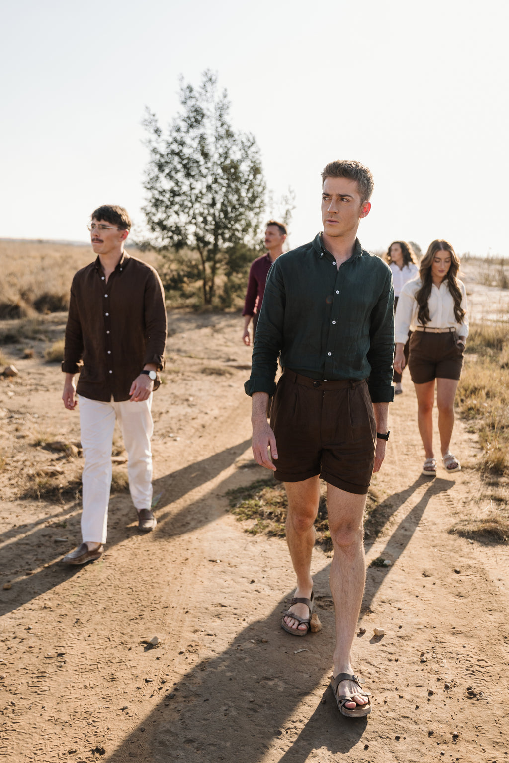 A group of five people walking outdoors on a dirt path in a sunny, dry landscape. The main subject in the foreground is a man wearing a Drakensberk dawn 100% certified Irish dark green linen in a long-sleeve button-up shirt and Lowveld Umber 100% European  brown linen shorts  with other men and women behind him in linen outdoor attire.