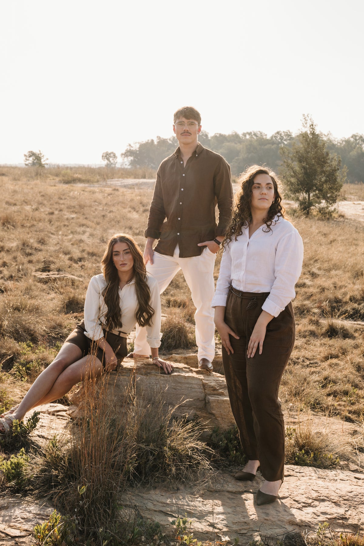 Three young adults posing outdoors in a dry grass field at sunset. The woman on the left sits on a rock wearing a cream Tsitsikamma Mist 100% Egyptian linen  button-down shirt Lowveld Umber 100% European brown linen shorts.