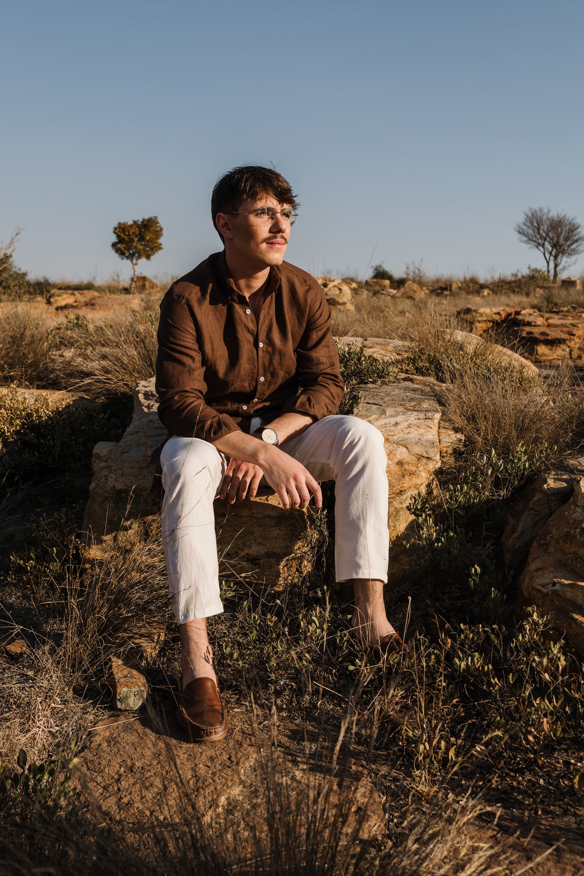 A man wearing a Lowveld Umber 100% European brown linen button-up shirt and cream Tsitsikamma Mist 100% Egyptian linen trousers siting  on a rock in a dry, rocky outdoor landscape under a clear blue sky.