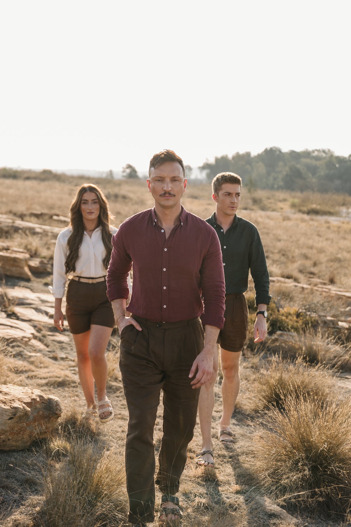 Three people walking outdoors in a grassy landscape during golden hour. The central person wears a Royal Karoo 100% certified Irish burgundy linen button-up shirt and Lowveld Umber 100% European dark brown trousers, while the person behind wears a Drakensberg dawn 100% certified Irish dark green linen shirt and Lowveld Umber 100% European brown linen shorts, and the person on the left wears a cream Tsitsikamma Mist 100% Egyptian linen  button-down shirt and Lowveld Umber 100% European brown linen shorts.