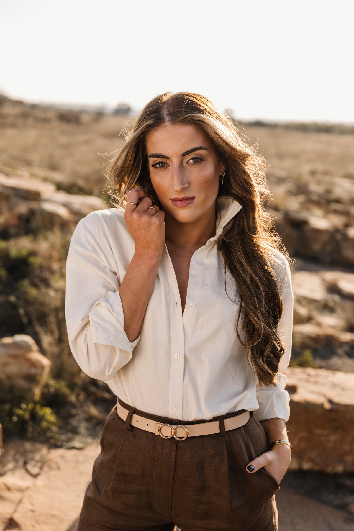 A woman with long wavy brown hair stands outdoors in a dry grassy field, wearing a cream Tsitsikamma Mist 100% Egyptian linen button-down shirt tucked into a Lowveld Umber 100% European brown linen  trousers with a tan belt.