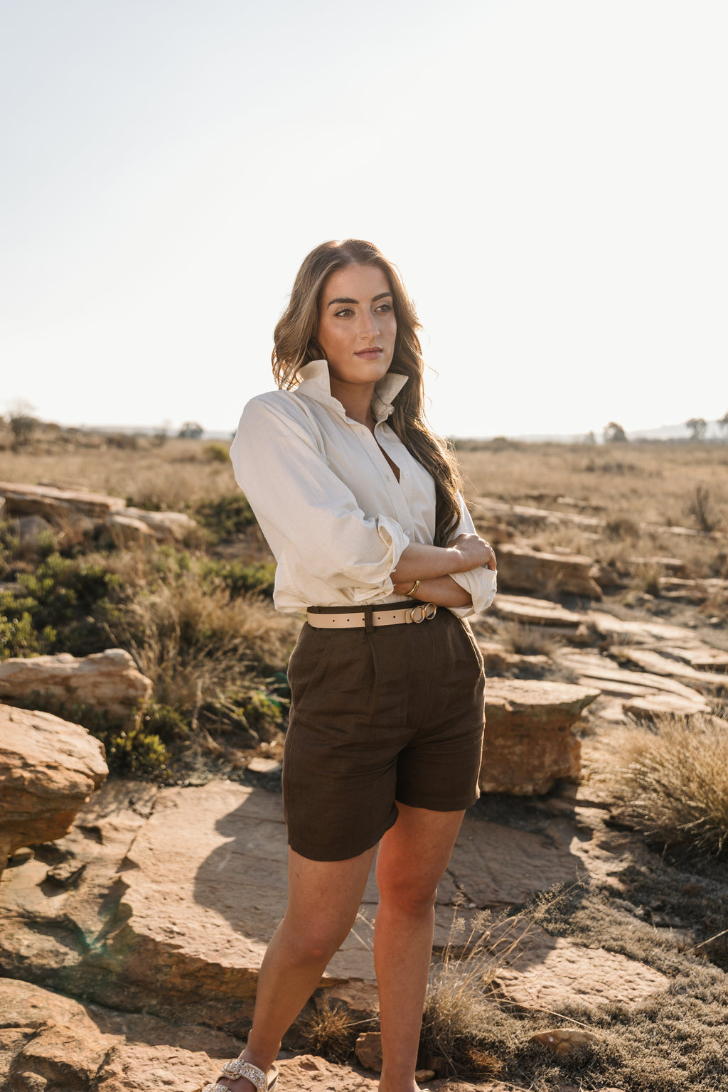 A woman stands outdoors in a rocky, dry landscape wearing a cream Tsitsikamma Mist 100% Egyptian linen button-up shirt and Lowveld Umber 100% European dark brown linen shorts.