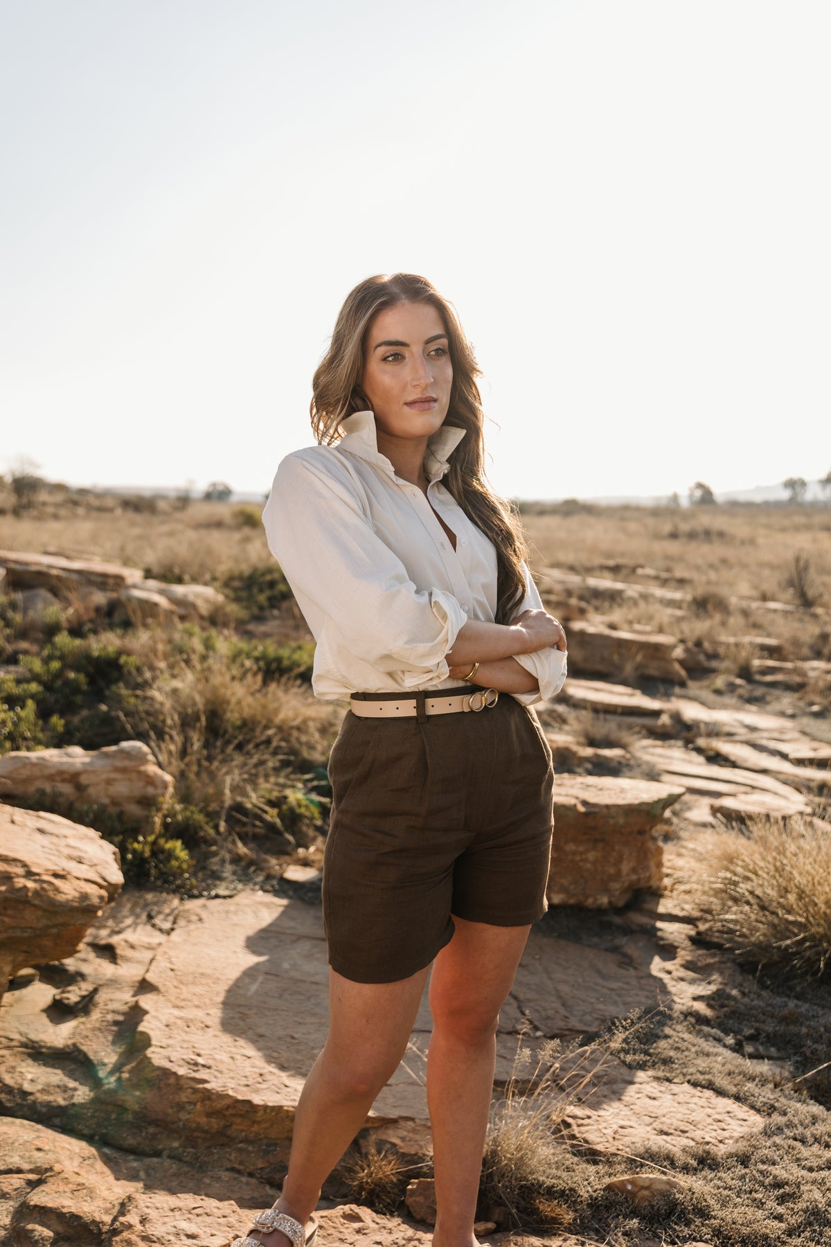 A woman stands outdoors in a rocky, dry landscape wearing a cream Tsitsikamma Mist 100% Egyptian linen button-up shirt and Lowveld Umber 100% European dark brown linen shorts.