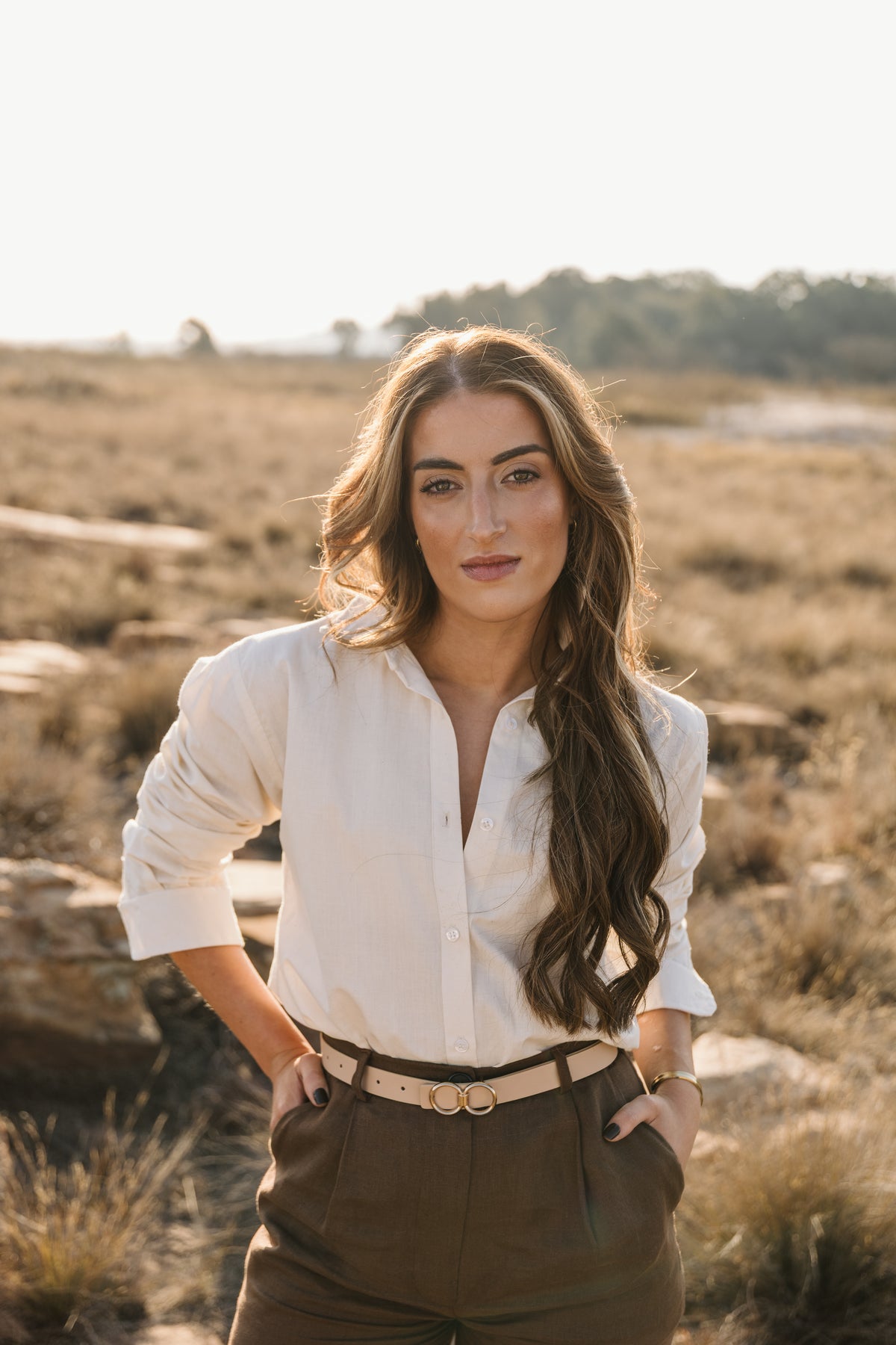 A woman with long wavy brown hair stands outdoors in a dry grassy field, wearing a cream Tsitsikamma Mist 100% Egyptian linen button-down shirt tucked into a Lowveld Umber 100% European brown linen trousers with a tan belt.